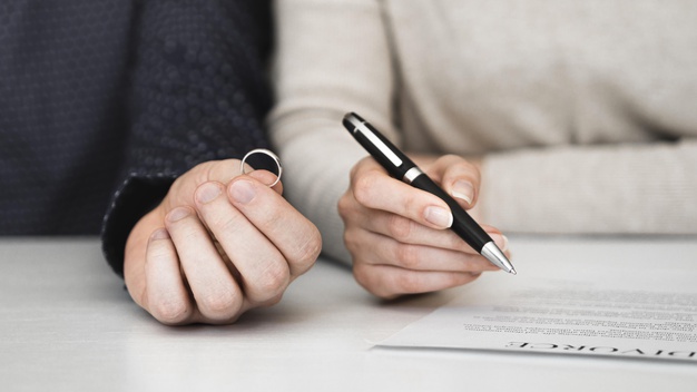Hand holding a ring next to another hand holding a pen poised above a divorce document.